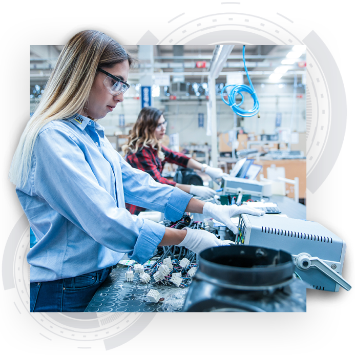 Ein Visual von zwei Frauen, die Elektroteile montieren in einer großen Produktionshalle. Im Hintergrund des Bildes sind zwei graue Designelemente in Form eines Präzisionsbauteils zu sehen.