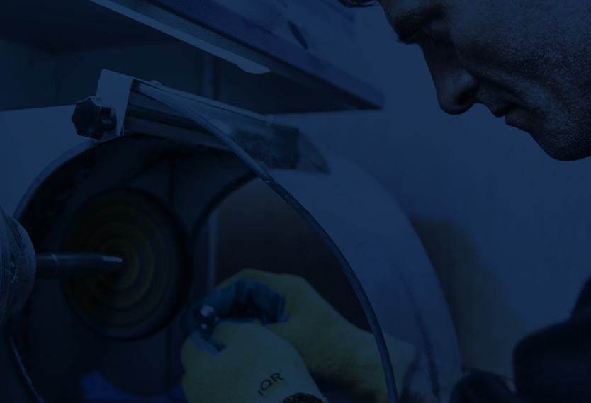 Ein Bildausschnitt im Querformat eines Mannes, der ein Metallwarenteil an einer Maschine nacharbeitet. Das Bild ist mit einem blauen Layer bearbeitet.